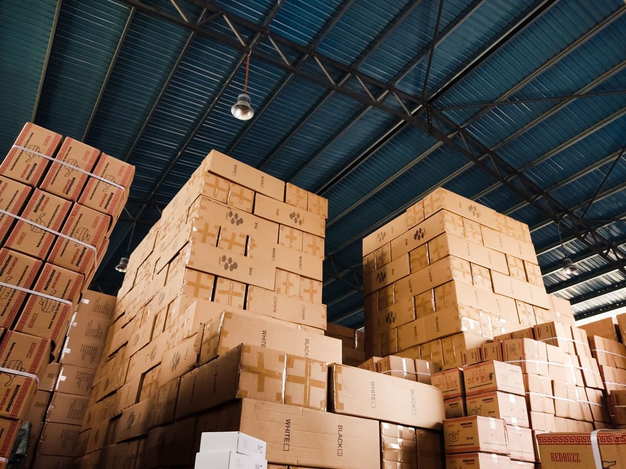 get-in-touch High stacks of cardboard boxes organized in a warehouse with a blue metal ceiling.