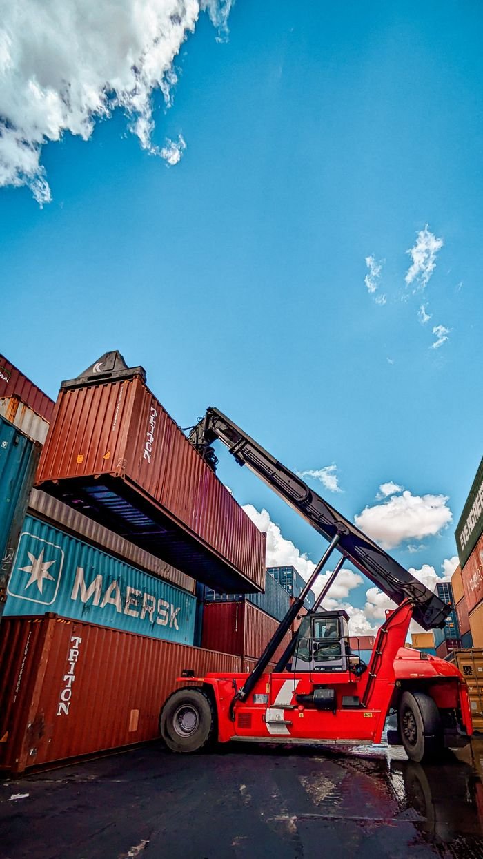 about-bg Red hydraulic crane handling shipping containers at a port under a clear blue sky.
